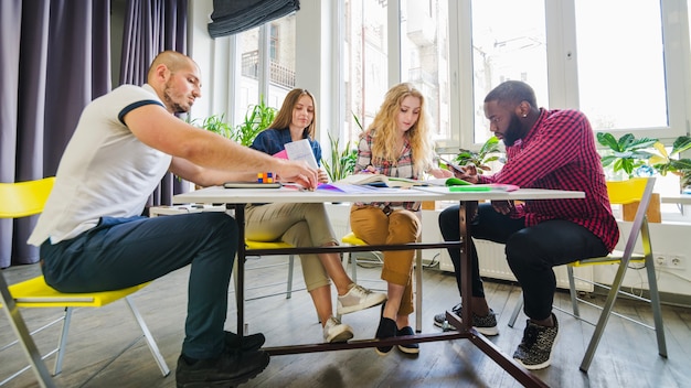 A group of students is gathered around a table, collaborating on a project. They are all looking at a tablet displaying research data, and they are discussing their findings. The scene emphasizes collaborative learning and the use of technology in education.