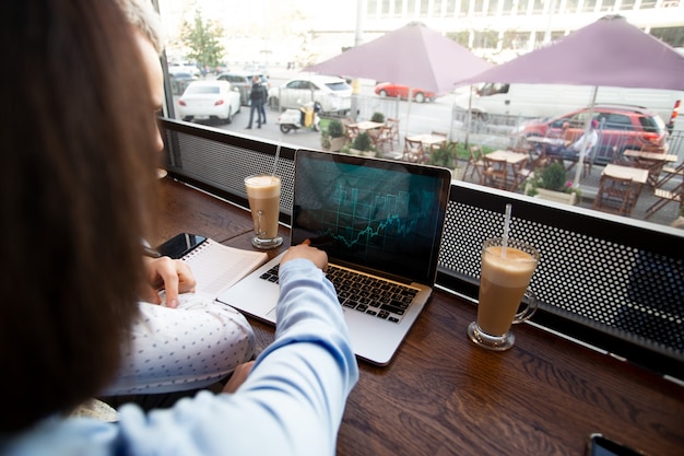 A person working on a laptop in a coffee shop, with various financial apps and charts displayed on the screen. The scene represents the flexibility and financial challenges of the gig economy.