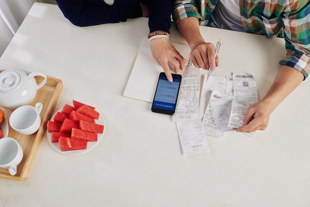 A person using a smartphone app to scan a receipt, with various other receipts scattered on a table. The background is a modern kitchen setting.