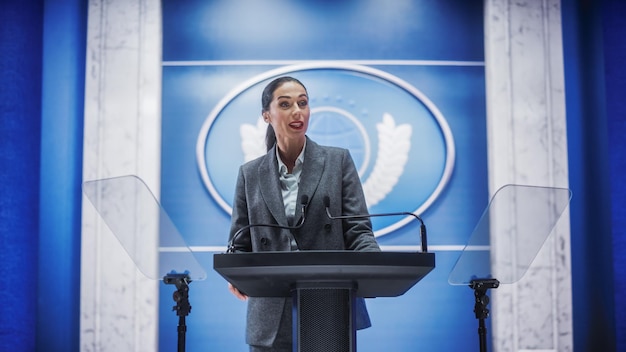 A person speaking at a podium during a conference. The person is energetic and enthusiastic, gesturing to the audience. The backdrop includes the company logo.
